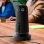 Owl Labs Meeting Owl 5 Pro 360 4K konferenču kamera, novietota vertikāli uz koka galda, ar vadības pogām un apļa logotipu, fona izplūdušas cilvēku figūras sanāksmes vidē.