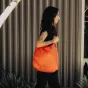 A person outdoors next to a vertical metal fence, wearing a black top and holding a bright orange Peak Design Packable Tote. The bag is partially open, revealing its interior, surrounded by green plants in the foreground and a b