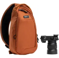 Think Tank TurnStyle 5L Urban Rust sling bag ar melnu sietu aizmuguri, ar rāvējslēdzēja galveno nodalījumu, planšetdatora kabatu un ūdens pudeles kabatu, blakus melnai kamerai ar Sony objektīvu.Think Tank TurnStyle 5L Urban Rust sling bag ar melnu sietu aizmuguri, ar rāvējslēdzēja galveno nodalījumu, planšetdatora kabatu un ūdens pudeles kabatu, blakus melnai kamerai ar Sony objektīvu.