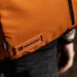 Крупный план ярко-оранжевого Wandrd PRVKE 21 Liter Sedona Orange Photo Bundle V3 с черной молнией, шестиугольным логотипом и названием бренда, тисненым на кожаной нашивке, носимого поверх черных брюк, с размытой рукой и браслето