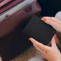 Melna Peak Design Passport Wallet turēts rokās ar oranžu nagu laku, blakus bordo čemodānam, izceļot tā plāno dizainu un magnētisko aizdari.