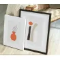 Nielsen baltās un melnās foto rāmji (15x20 cm) ar dekoratīviem plakātiem, novietoti uz koka grīdas blakus austa krēsla un adīta segu, parādot minimalistisku oranžas vases ilustrāciju un abstraktu ģeometrisko dizainu.