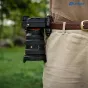 Leofoto QF-03 + QP-38 black backpack quick‑release mount attached to a belt, featuring automatic locking, silicone pad and Arca‑Swiss compatibility, with a camera on a person's waist in an outdoor setting.